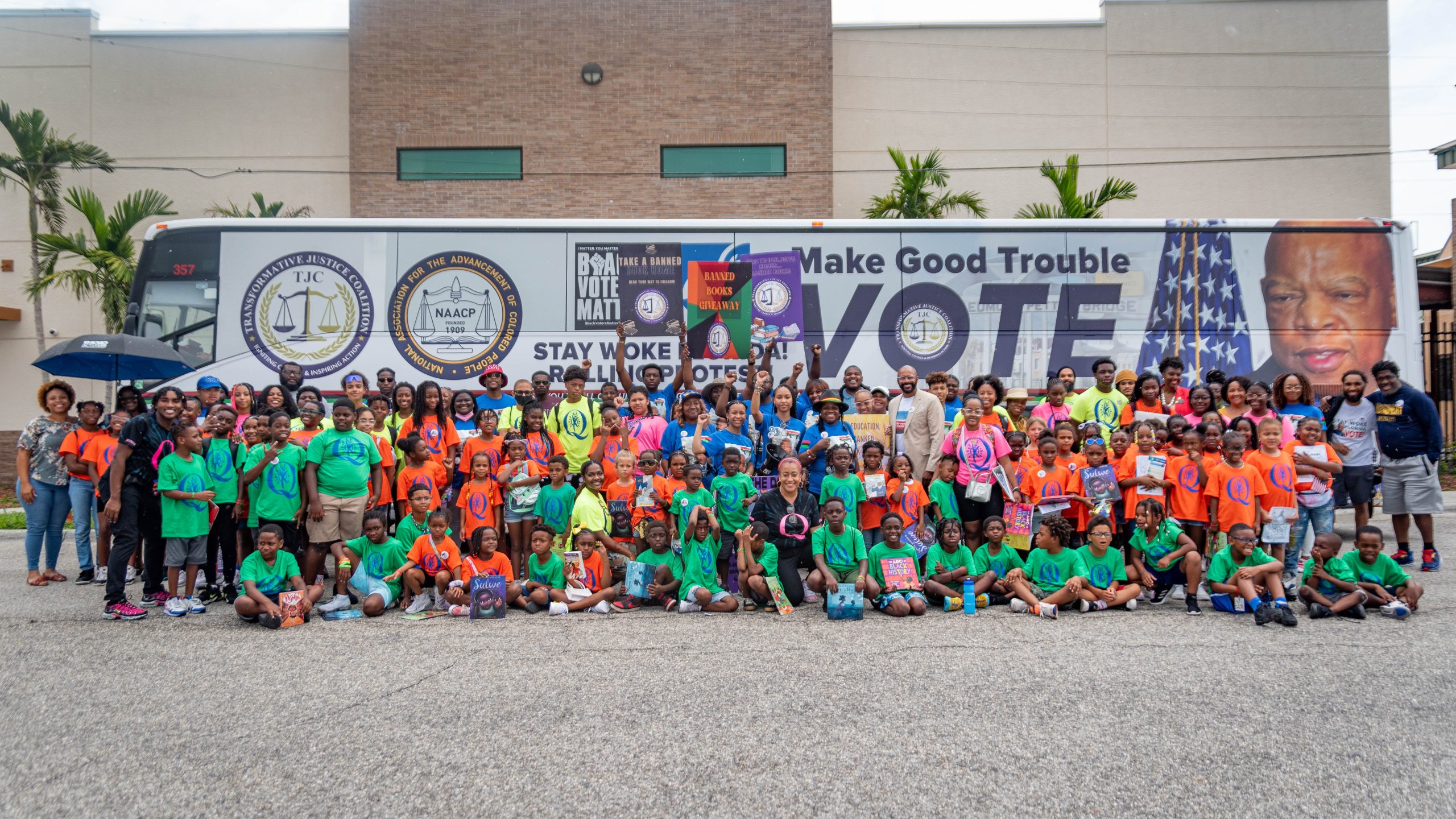A large group of children and adults gathers in front of a bus promoting voting and literacy, holding books and signs for a "Banned Books Giveaway."
