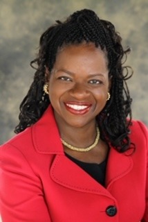 Smiling woman with braided hair wearing a red blazer and pearl necklace, set against a blurred neutral background.