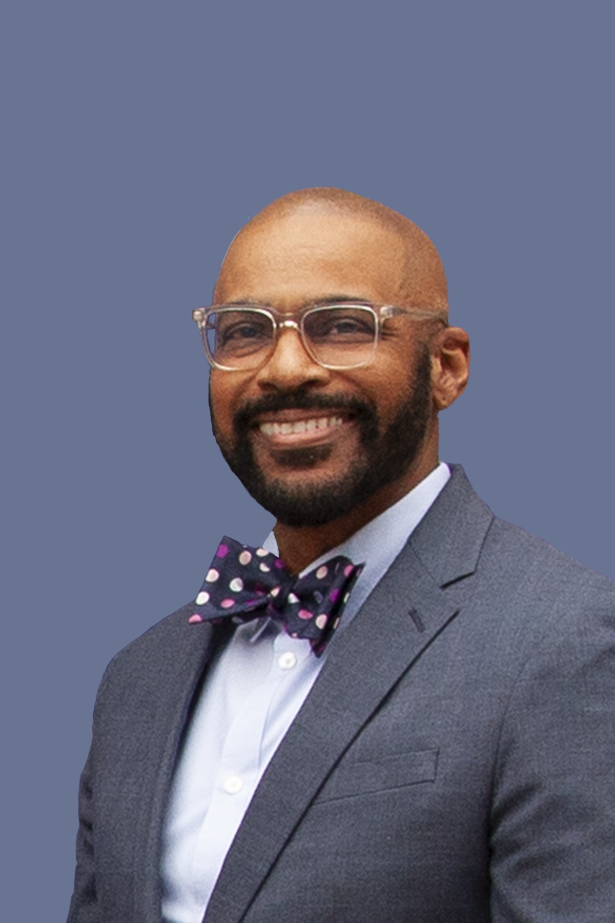 Professional portrait of a man in a gray suit and colorful polka dot bow tie, smiling against a muted blue background, exuding confidence.