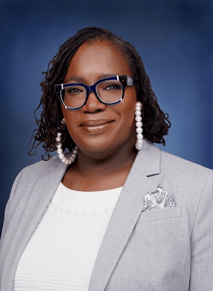 A woman with curly hair, wearing glasses and large pearl earrings, smiles confidently in a light grey suit against a blue backdrop.