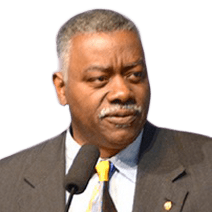 A man with short gray hair speaks into a microphone, wearing a suit and a tie. His expression is serious, indicating he is addressing an important topic.